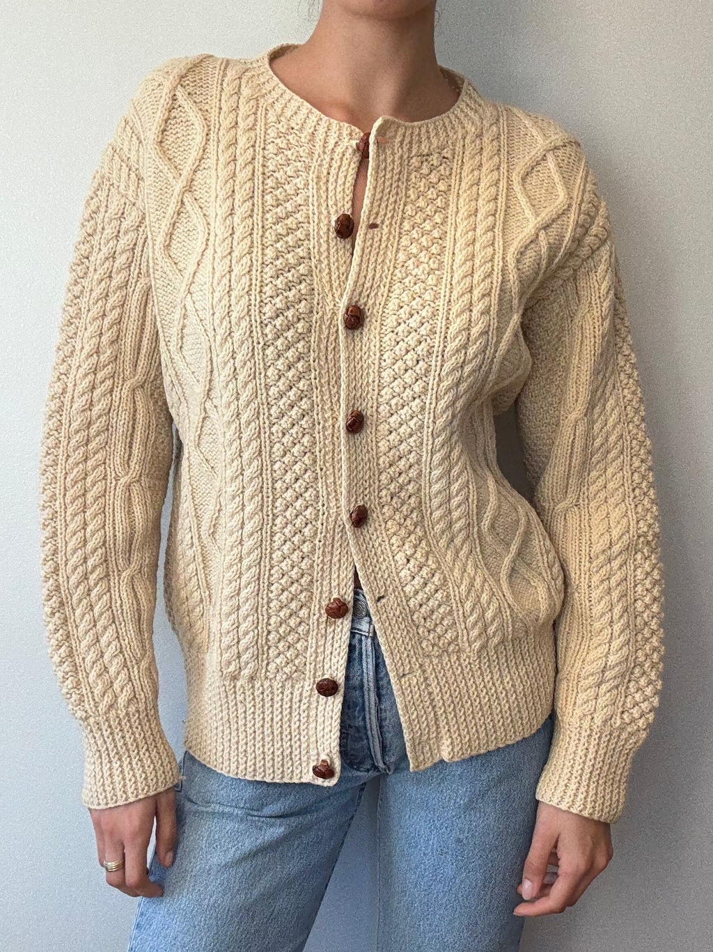 Person wearing a beige cable knit cardigan with brown buttons against a plain background