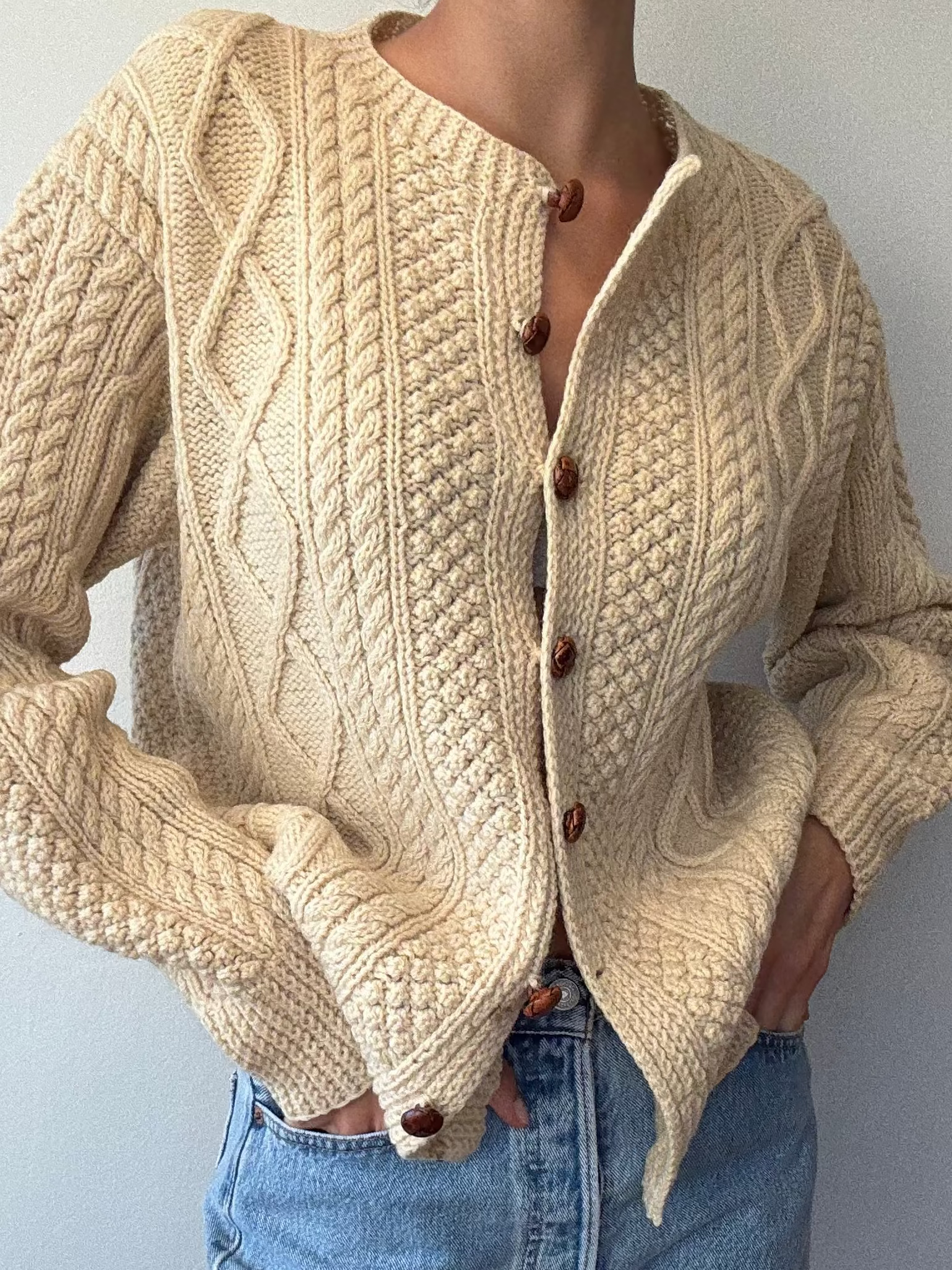 Person wearing a beige knitted cardigan and blue jeans against a plain background