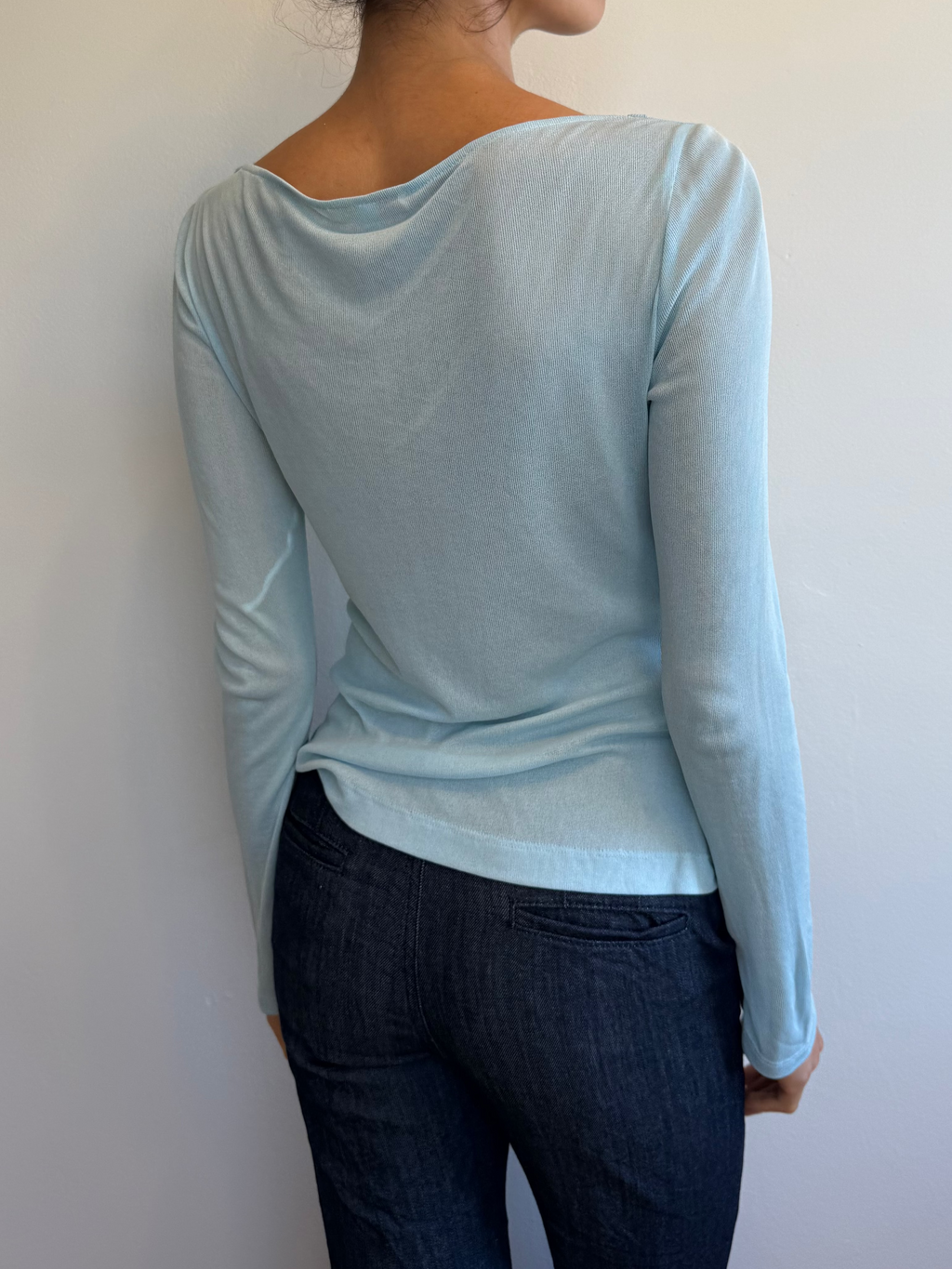 Woman wearing a light blue long-sleeve top and dark jeans against a white wall