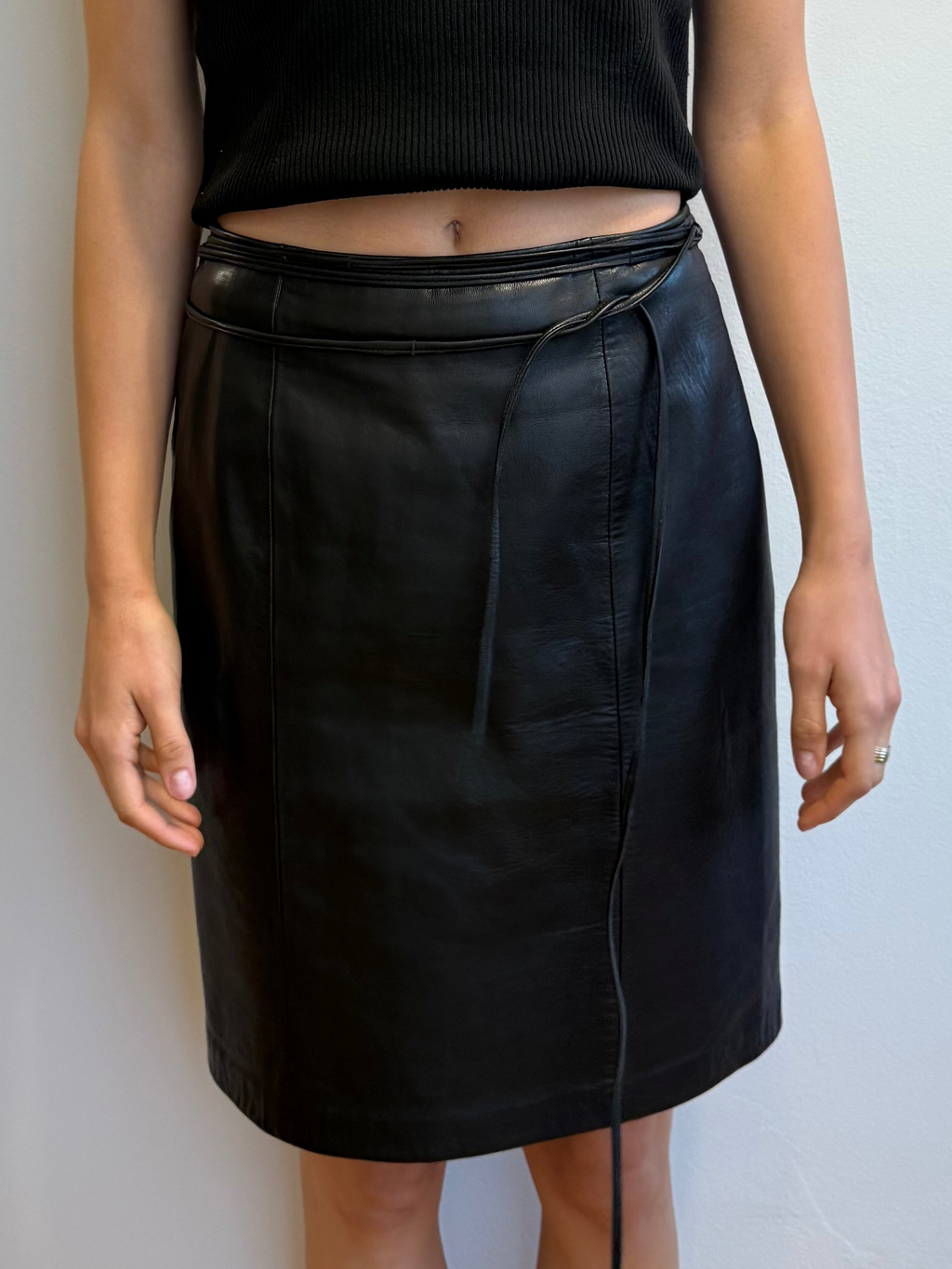 Person wearing a black sleeveless top and black leather skirt against a plain wall.