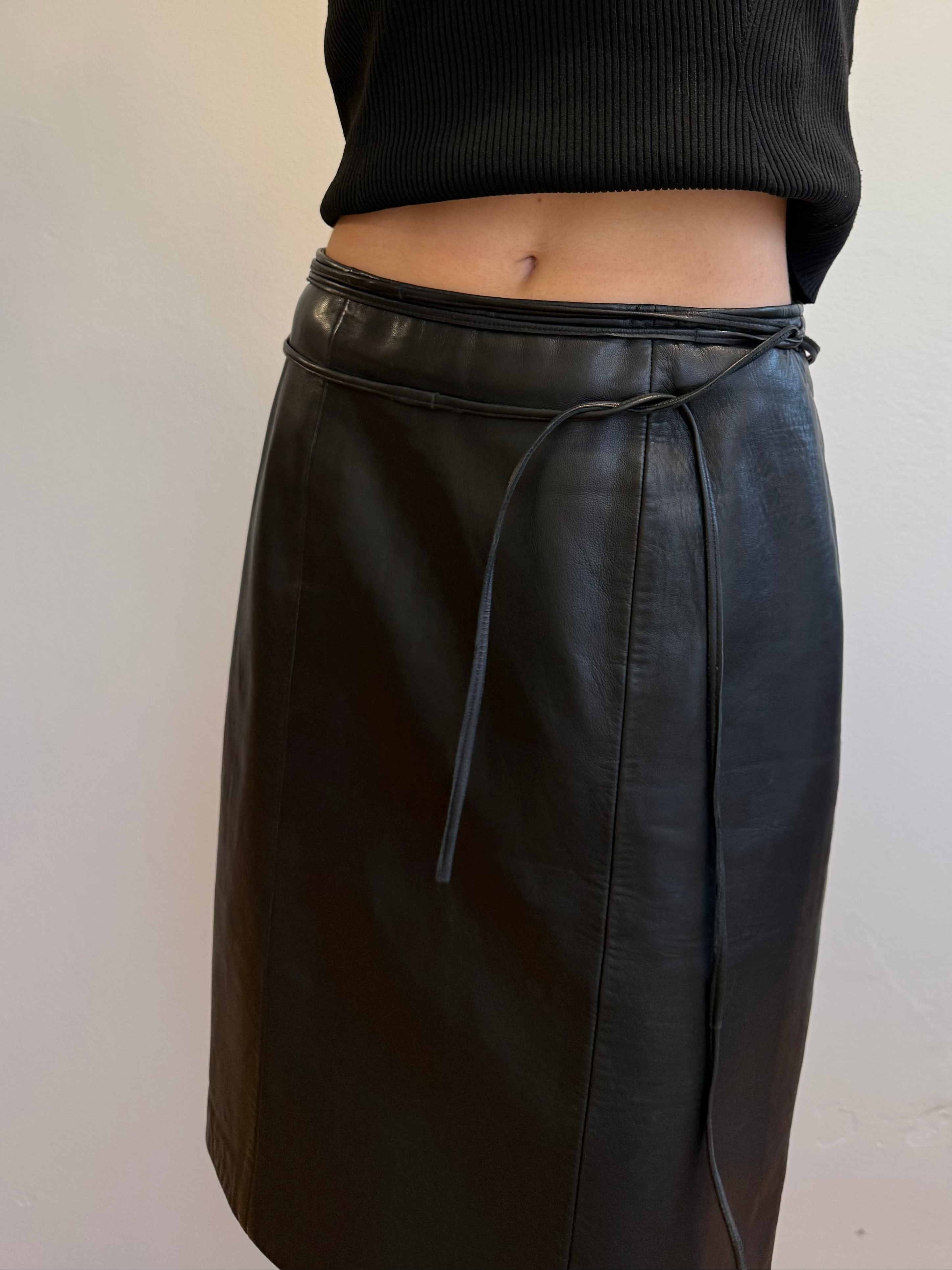 Person wearing a black sleeveless top and black leather skirt against a plain wall.