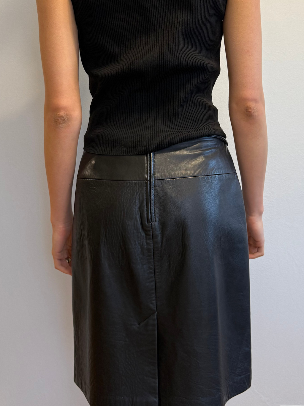 Person wearing a black sleeveless top and black leather skirt against a plain wall.