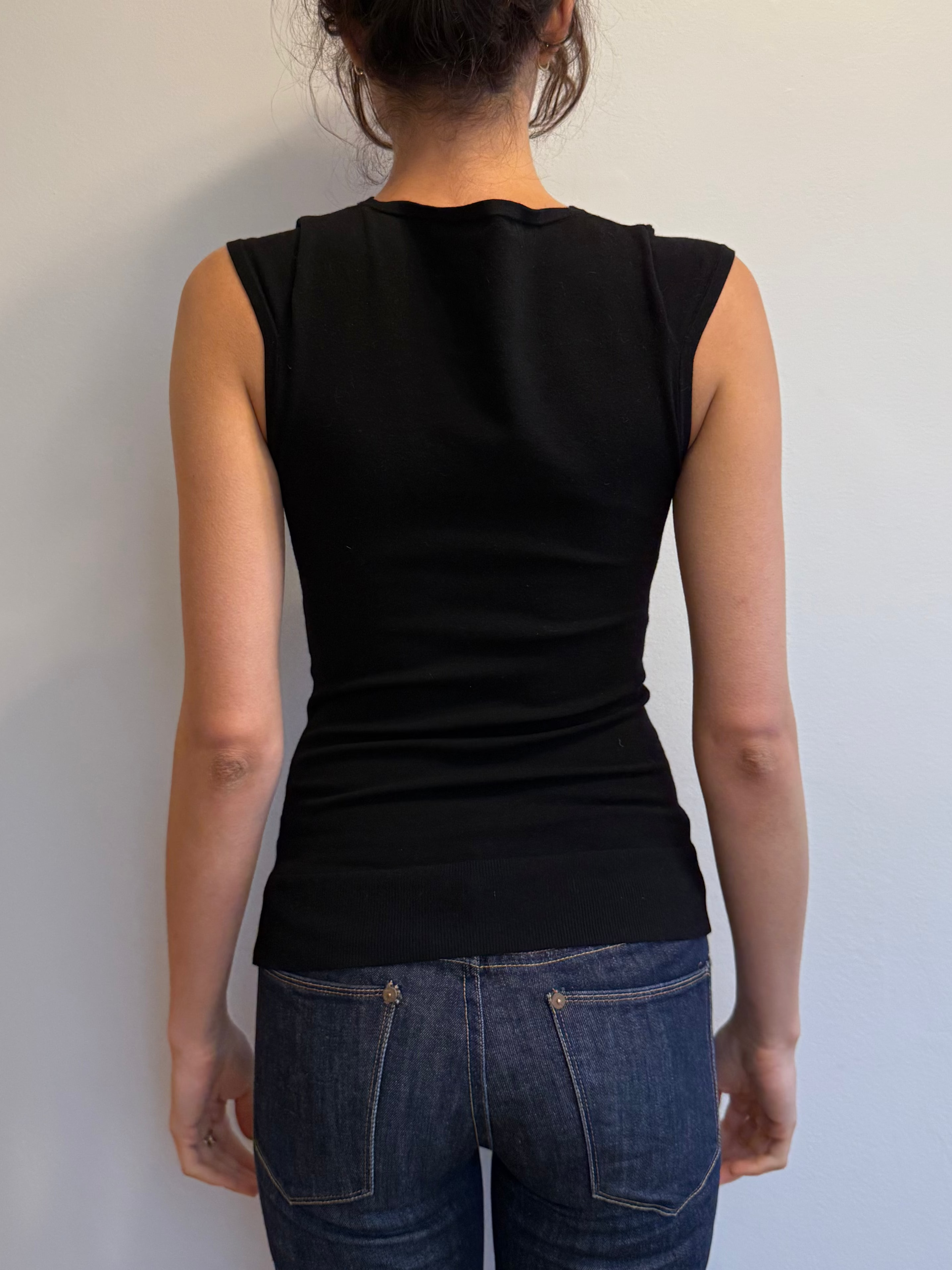 Person wearing a black sleeveless top and blue jeans against a plain white wall