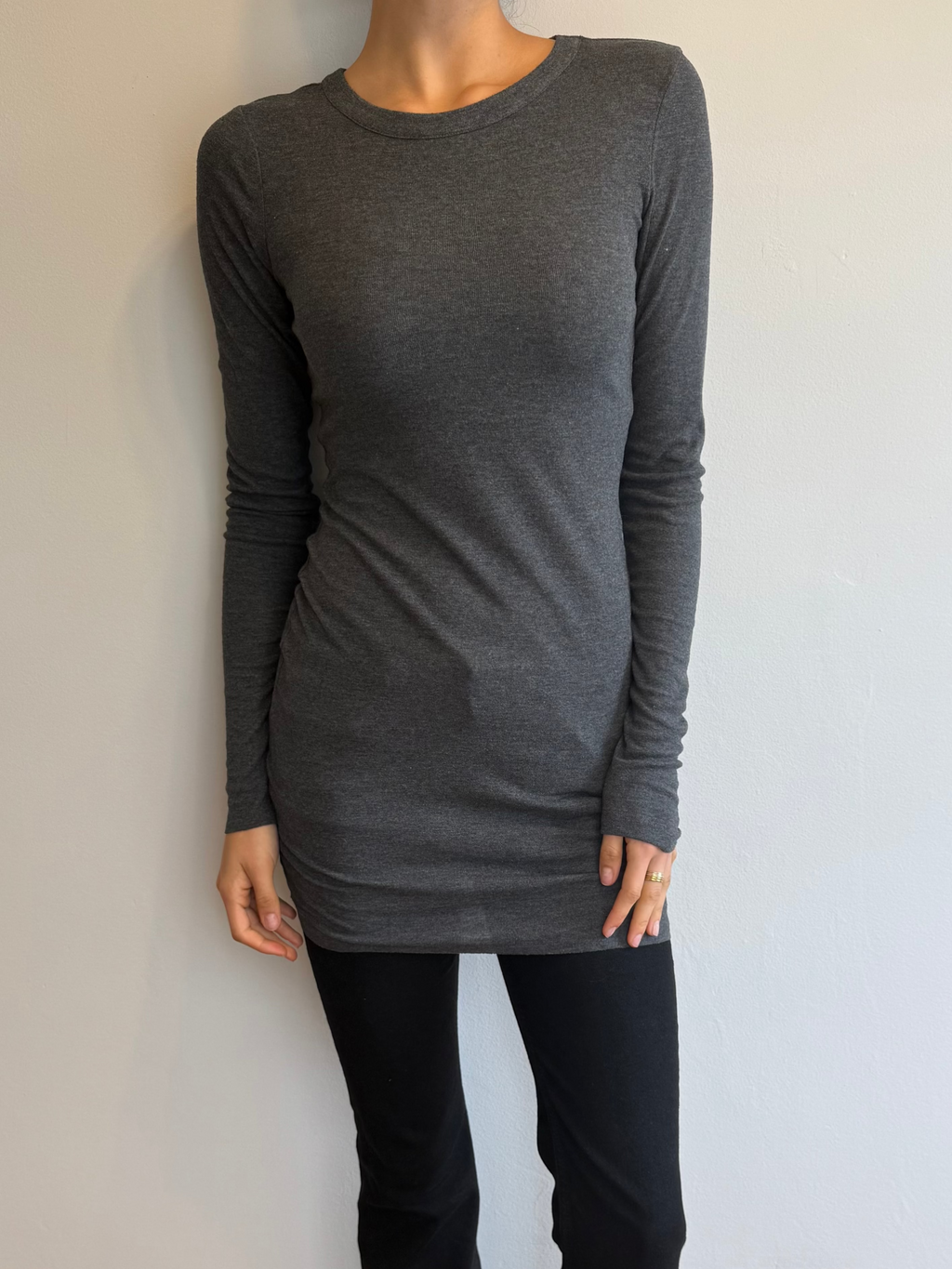 Person wearing a gray long-sleeve top and black pants against a plain wall.