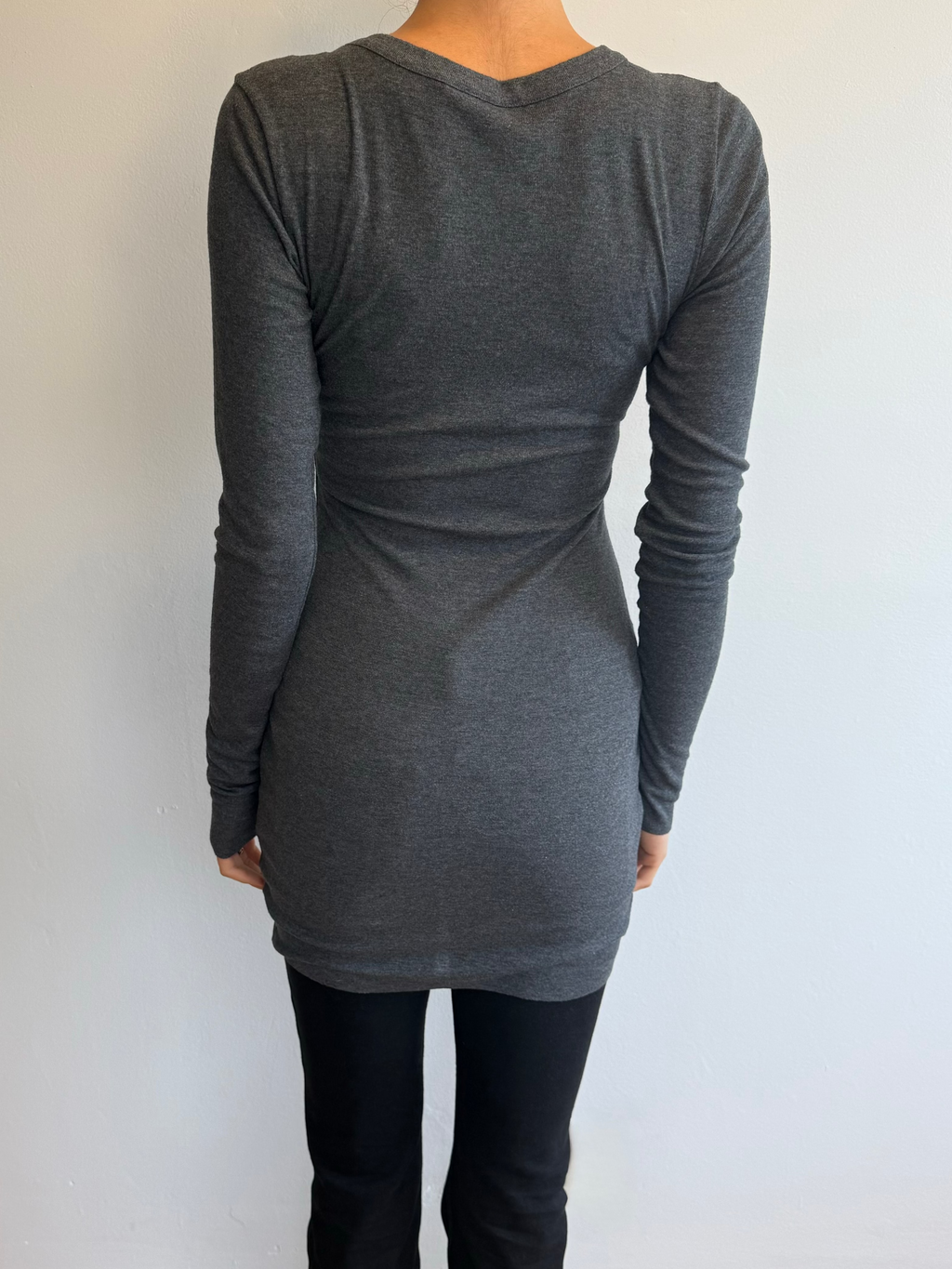 Person wearing a gray long-sleeve top and black pants against a white wall.