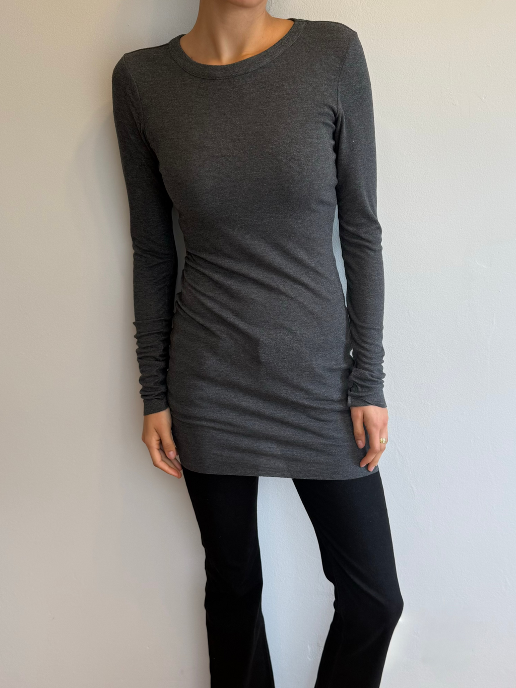 Person wearing a gray long-sleeve top and black pants against a plain white wall.